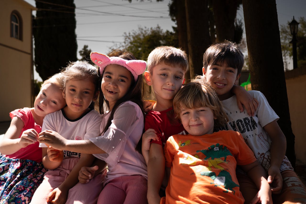 A diverse group of cheerful children hugging and smiling in an outdoor setting under sunlight.